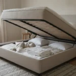 Chambre scandinave avec lit coffre ouvert sur des vérins, linge de lit plié dans le coffre, tête de lit en lin et lumière naturelle