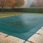 Piscine enterrée couverte d'une bâche d'hivernage vert foncé bien tendue, feuilles mortes sur la margelle en pierre, jardin d'automne dans la brume du matin